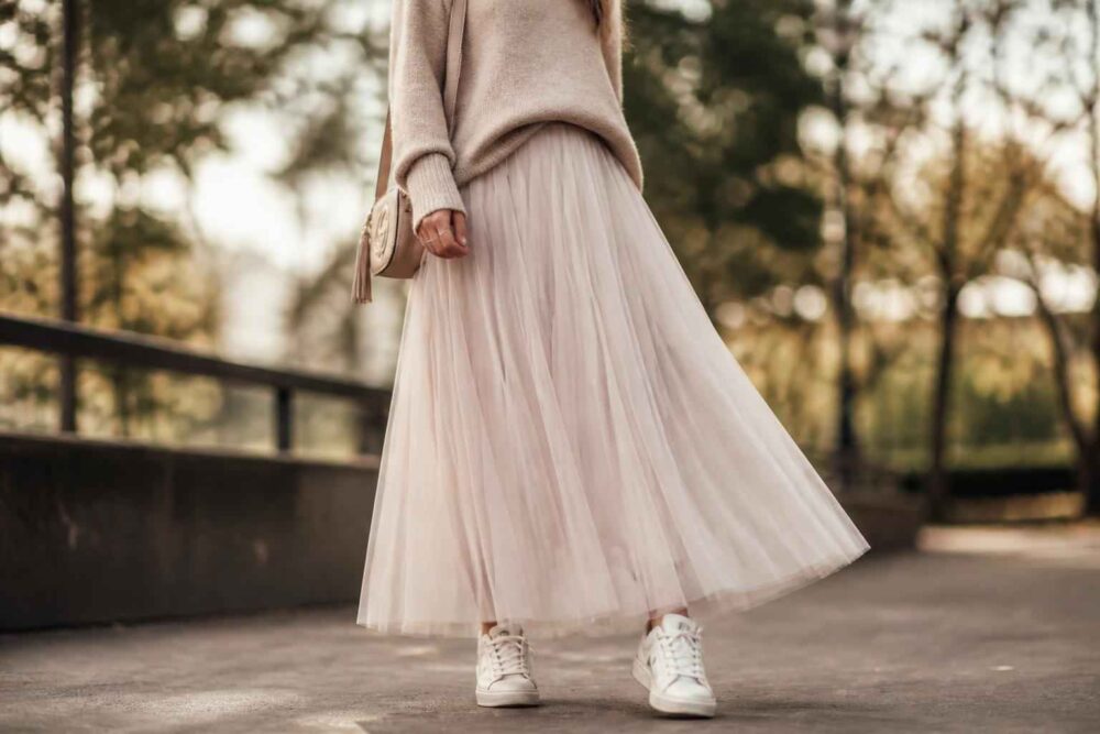 Kobieta w beżowym swetrze i długiej plisowanej spódnicy spaceruje w parku, ubrana w białe sneakersy